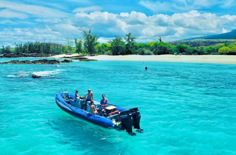People on a small motorboat in clear blue water near a lush green island with a sandy beach.